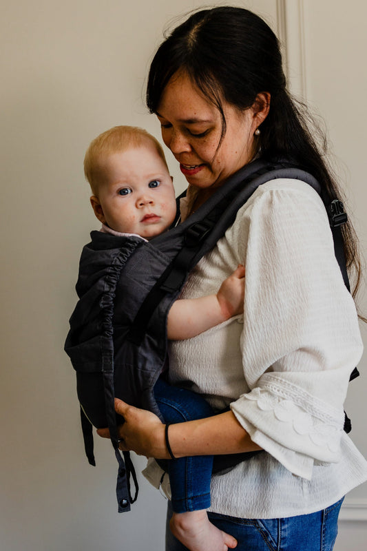 Moeder draagt haar baby comfortabel op de buik in een ergonomische Juami draagzak, terwijl ze rustig binnen is, omringd door een warme en gezellige sfeer.