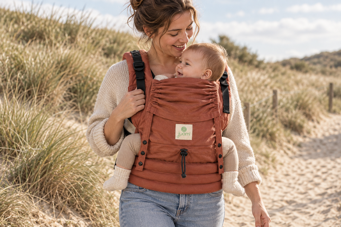 Moeder wandelt met haar baby in een terracotta draagzak langs een duinpad.