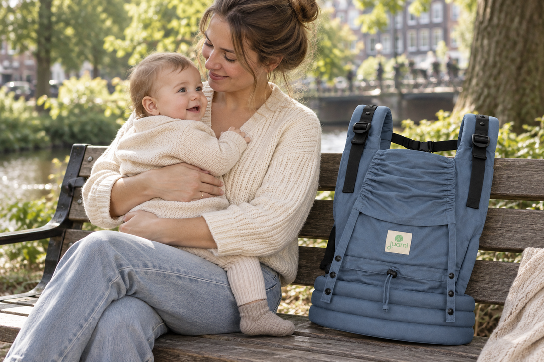 Moeder neemt een pauze op een bankje met haar baby op schoot en een blauwe draagzak naast zich.