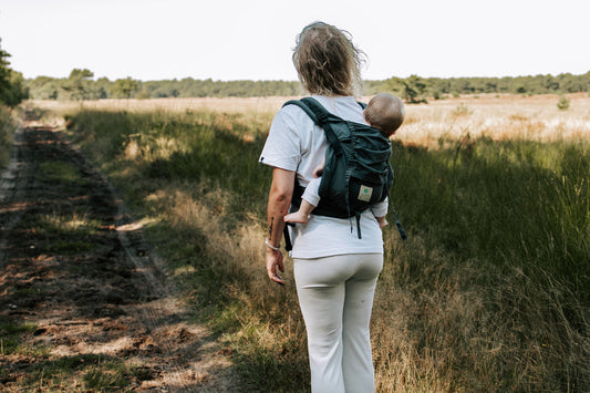 Moeder draagt haar baby op de rug in een ergonomische Juami draagzak tijdens een wandeling in het bos.
